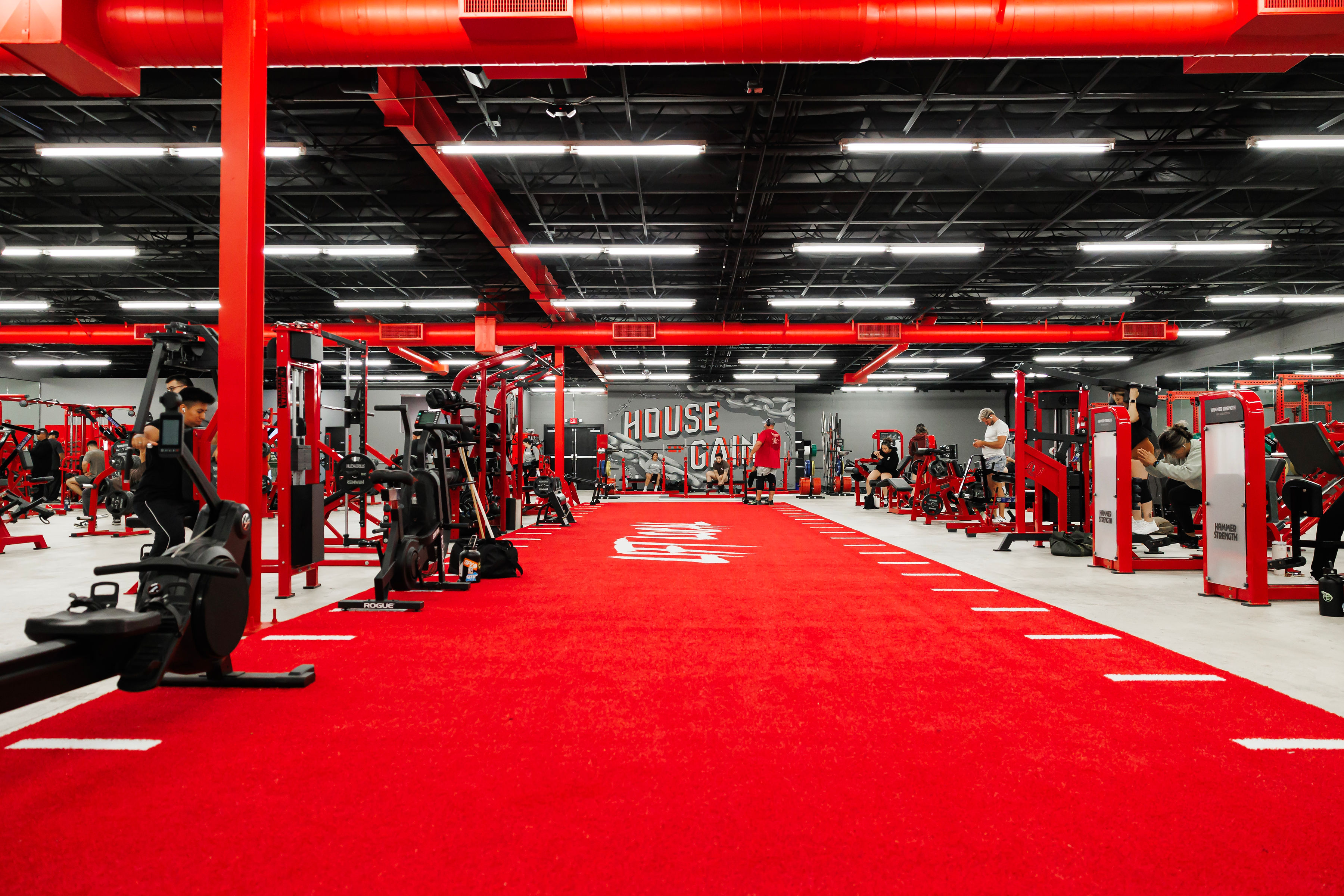 Wide view of the House of Gains floor — red racks and machines flanking the red turf runway, with the HOUSE OF GAINS mural on the back wall